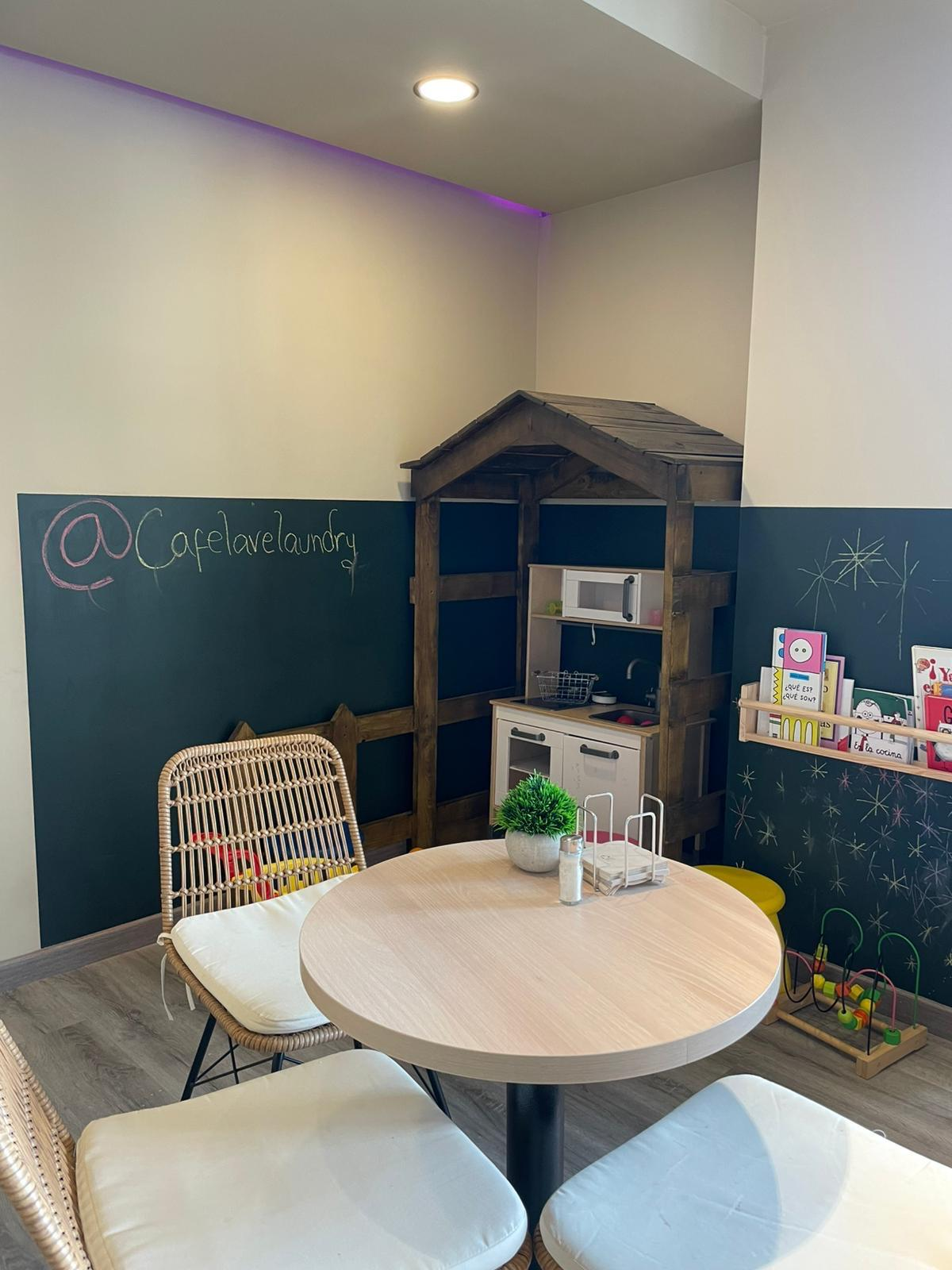 Rincón infantil dentro de una cafetería con una mesa redonda, sillas de mimbre, una casita de madera, cocina de juguete, estante con libros y juegos didácticos sobre el suelo.