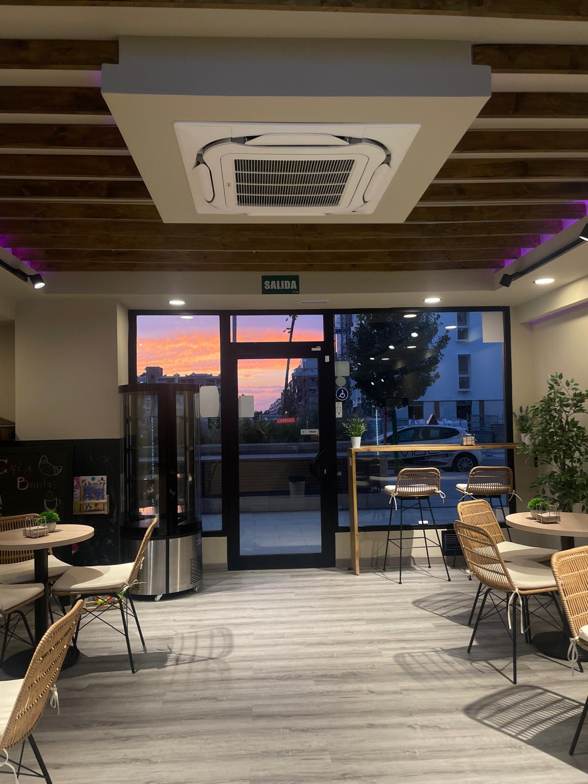 Interior de una cafetería con piso gris, techo con vigas de madera y aire acondicionado empotrado. Hay mesas con sillas de mimbre y una barra alta junto a la puerta de vidrio, por la que se ve un atardecer color naranja y rosa.